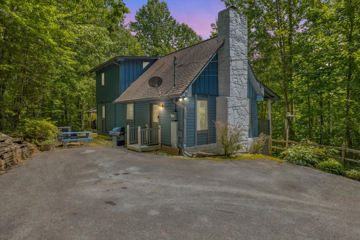 The image showcases the inviting exterior of a charming blue cabin, accented by a rustic stone chimney, nestled among lush trees. A spacious deck offers spots for outdoor dining and relaxation, while a cozy fire pit area invites guests to gather and unwind against a tranquil forest backdrop.
