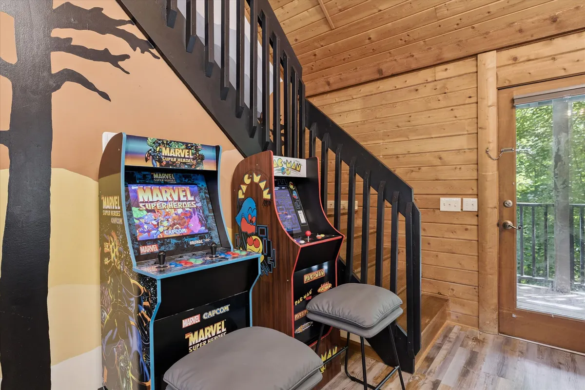This image highlights the arcade area inside the cabin. It features classic arcade machines next to a wooden staircase and large windows, inviting both nostalgic fun and social connection. There are visible cues of people playing and enjoying snacks.