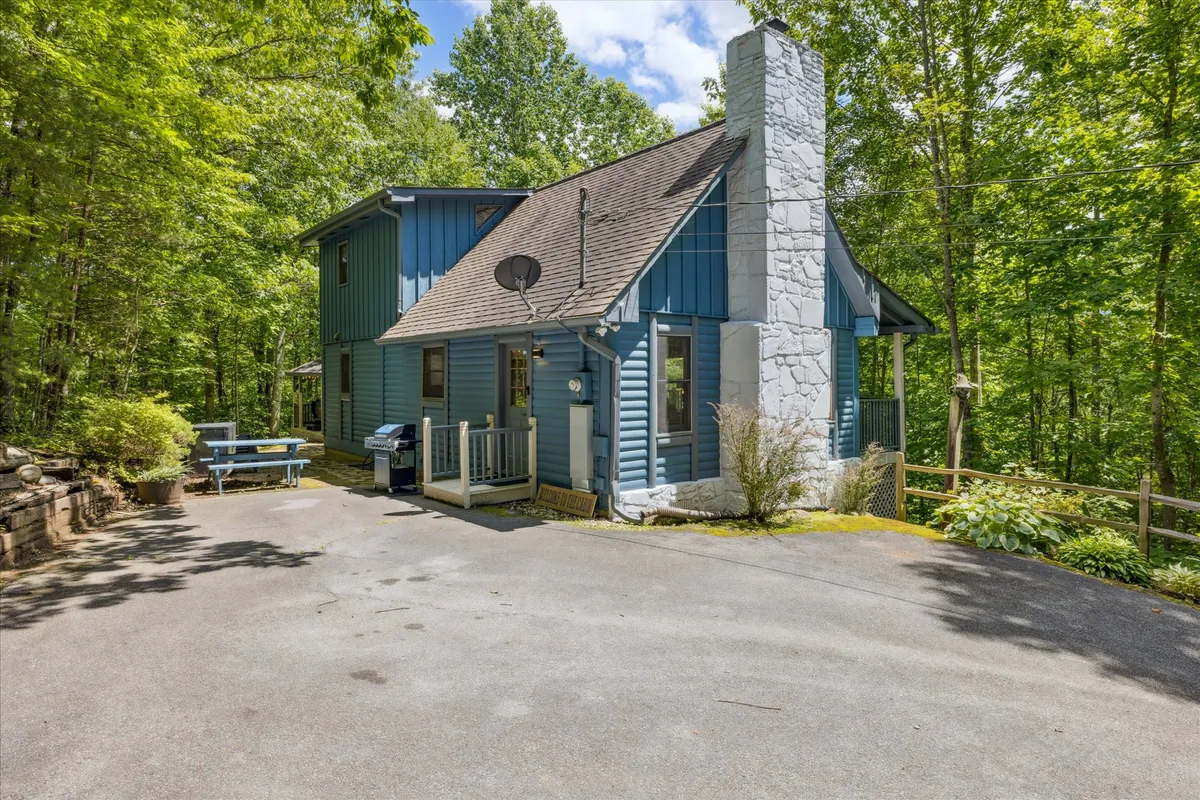 This exterior image showcases the blue exterior of the house, a stone chimney, the deck area with comfortable seating, and surrounding lush greenery. The deck is oriented to take full advantage of sunset views, inviting outdoor relaxation and gatherings.