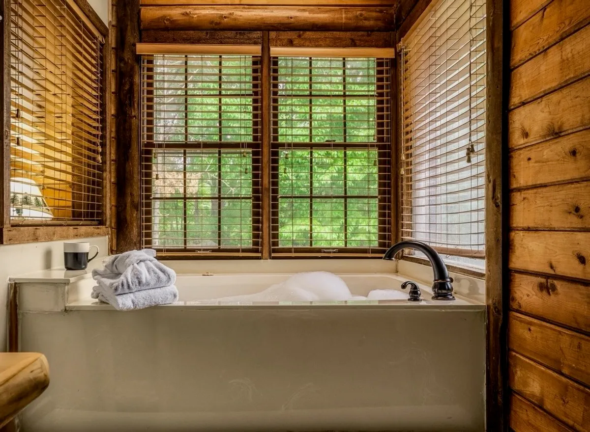 A serene bathroom with a deep soaking bathtub set against wooden walls and windows that let in gentle light. Towels and a cup on the tub’s edge create an inviting scene perfect for relaxation or self-care. The mood is quiet and spa-like.
