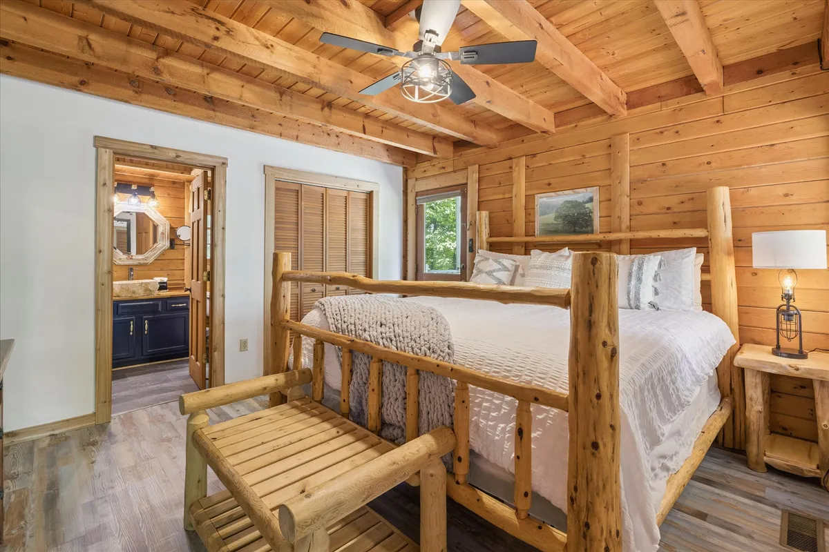 A rustic bedroom scene featuring a log bed frame topped with warm blankets and pillows, a ceiling fan overhead, a wooden bench at the foot of the bed, and natural wooden walls. A decorative lamp adds a soft, intimate glow to the room.