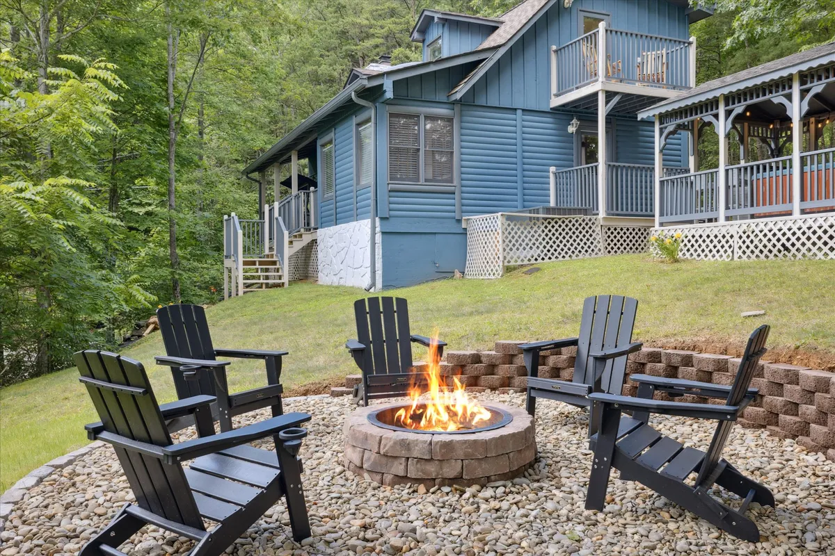 A cozy fire pit surrounded by Adirondack chairs sits in a grassy yard beside the blue house, backed by verdant trees. The setup invites friends to gather, share stories, and enjoy evenings under the stars.