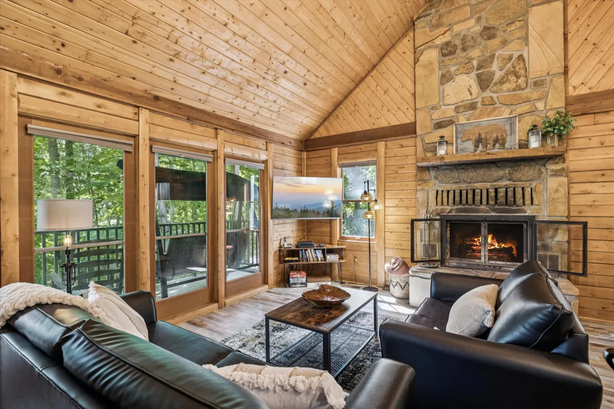 This image showcases a welcoming living room with a prominent stone fireplace, large windows letting in natural light, comfortable seating, and wooden beams. The setup encourages relaxation, socializing, and warmth, aligning with the communal, cozy atmosphere described in the section.