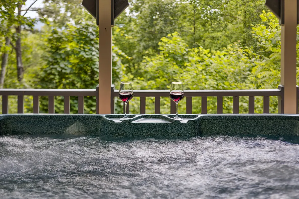 An inviting outdoor hot tub nestled among lush greenery, with wine glasses resting on the edge. The scene suggests relaxation, romance, and the enjoyment of mountain air and views, particularly at sunset or under the stars.