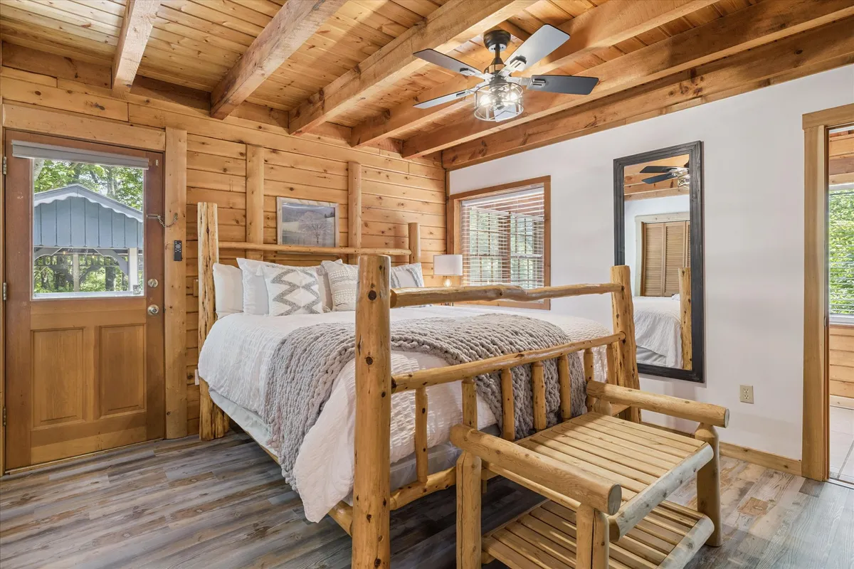This image features a cozy bedroom with a log bed, a wooden chair beside the window, and soft lighting streaming in. The window frames a peaceful view of nature, enhancing the rustic, tranquil ambiance. The bedding looks plush and inviting, and the decor subtly incorporates natural, mountain-inspired elements.